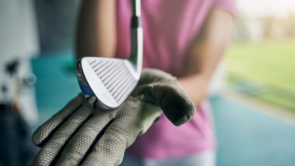 A person in a pink shirt grips a golf club with a gloved hand, the focus on the polished club head, reflecting how to clean golf irons to perfection. The background is softly blurred, emphasizing the pristine condition achieved without a golf club cleaner.